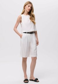 Bílý bezrukávový crop top spárovaný s bílými šortkami s vysokým pasem, zdobený černým opaskem, a černými skluznými sandály s uzlovým detailem.
