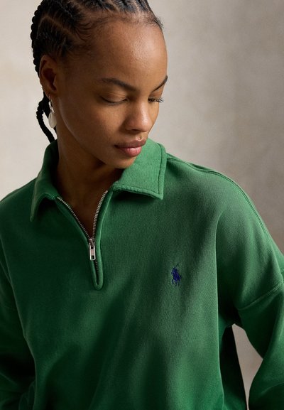 Femme aux cheveux tressés portant un pull zippé vert avec col et petit logo brodé, regardant vers le bas sur un fond neutre.