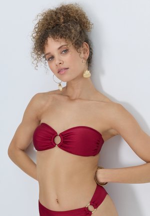 Woman with curly hair wearing a strapless red bikini with ring details, gold seashell earrings, and a gold bracelet, posing against a white background.