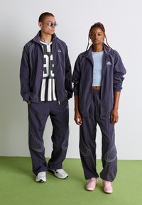 Két modell azonos sötétlila Adidas tréningruhát visel; a férfi napszemüveget és fekete-fehér mez visel, míg a nő világoskék trikót öltött magára.
