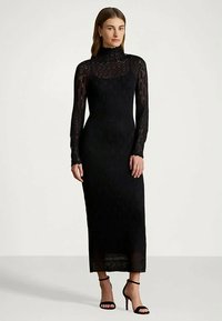 Mulher vestindo um vestido longo justo de renda preta com gola alta e mangas longas, combinado com sandálias de salto alto pretas, em pé contra um fundo branco.