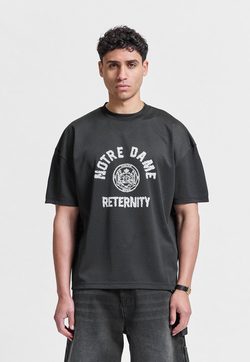 Jeune homme aux cheveux courts et bouclés portant un T-shirt en mesh noir avec le texte et l'emblème « NOTRE DAME ETERNITY », debout devant un fond uni.