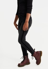 Úzké černé džíny s hladkou texturou, vysokým pasem a bočními pruhy, spárované s vínovými šněrovacími botami a přiléhavým černým topem s dlouhým rukávem.