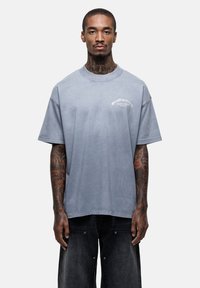 Szara, luźna koszulka z miękkiego materiału, z okrągłym dekoltem i krótkimi rękawami. Na przodzie nadrukowane logo tekstowe.