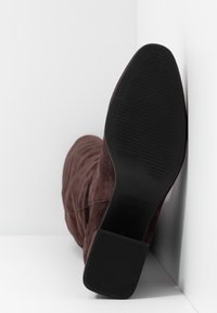 Bottes hautes en daim marron, avec une texture lisse et une semelle en caoutchouc noir plate. Le talon est carré et possède une surface texturée pour une meilleure adhérence.