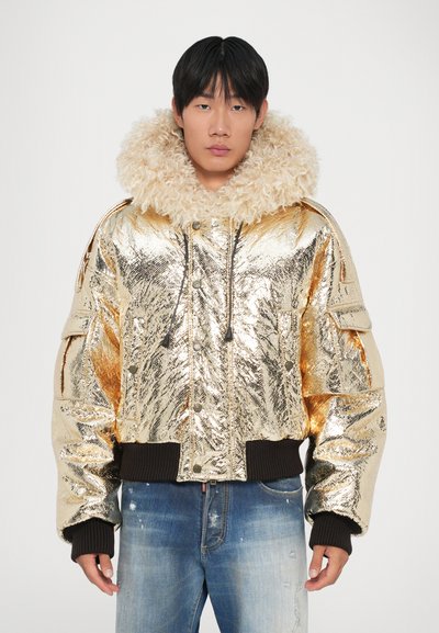 Veste bomber métallique dorée avec un col en fourrure beige et duveteux, des poignets côtelés et une fermeture éclair sur le devant. Le mannequin porte un jeans en denim usé.