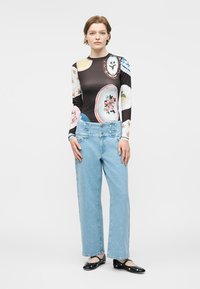 Kvinde står iført en langærmet top med mønster, lyseblå højttaljede jeans og sorte flade sko med ankelremme.