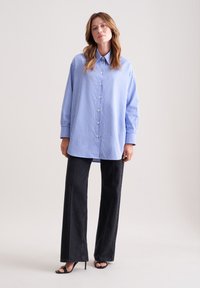 Camicia oversize a strisce blu chiaro e bianche con bottoni frontali, maniche lunghe e pantaloni larghi neri in denim. Tacchi aperti neri.