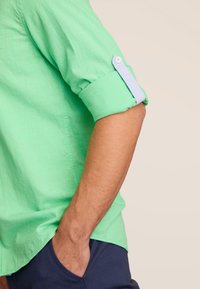 Lichtgroen shirt met opgerolde mouwen en een knoopsluiting. De stof lijkt textuur te hebben. Gdragend met donkerblauwe broek.