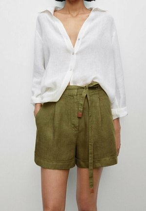Kvinde iført en løs hvid skjorte med knapper og opkrøllede ærmer og højtaljede olivengrønne shorts bundet med bælte.