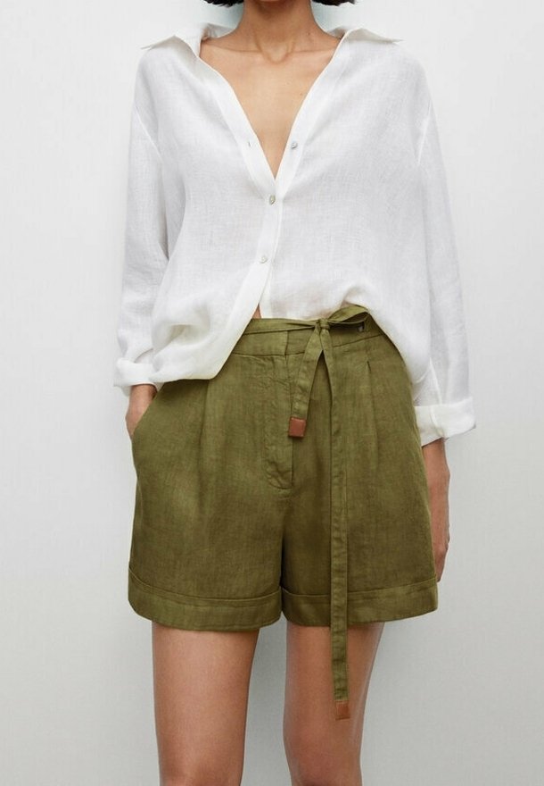 Femme portant une chemise blanche ample boutonnée avec les manches retroussées et un short taille haute vert olive noué avec une ceinture.