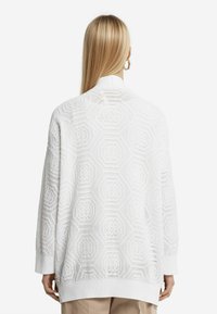 Vit stickad tröja med ett öppet hexagonmönster, långa ärmar och ribbade manschetter, i en avslappnad silhuett utan synliga knäppningar.