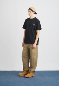 Camiseta negra de algodón con un gráfico en la parte frontal, pantalones anchos color caqui y botas de cordones color marrón claro. Gorra beige, llevada hacia atrás.