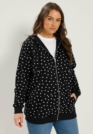 Kvinde iført en sort hættetrøje med hvidt polkaprikket mønster, en hvid skjorte under og blå jeans mod en ensfarvet baggrund.
