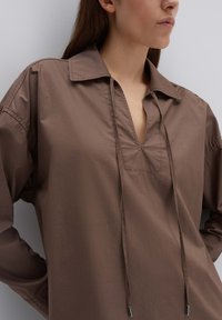 Bruine katoenen blouse met een kraag, V-vormige halslijn en trekkoorddetail. Heeft lange mouwen en een gladde, licht glanzende textuur.