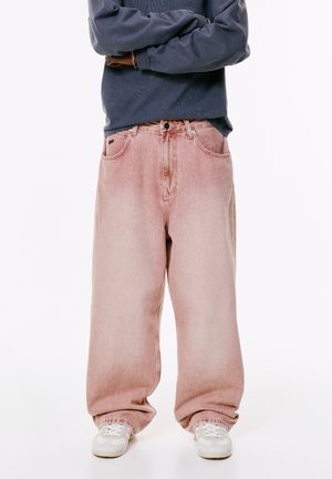 Person wearing oversized faded pink jeans with loose cuffs and white sneakers, standing with arms crossed against a white background.
