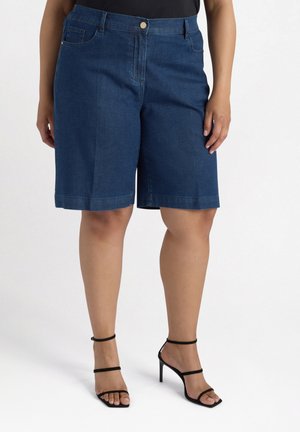 Bermudas de denim en azul oscuro, con un bolsillo frontal, corte recto y dobladillo justo por encima de la rodilla. Combinadas con tacones negros con tiras.