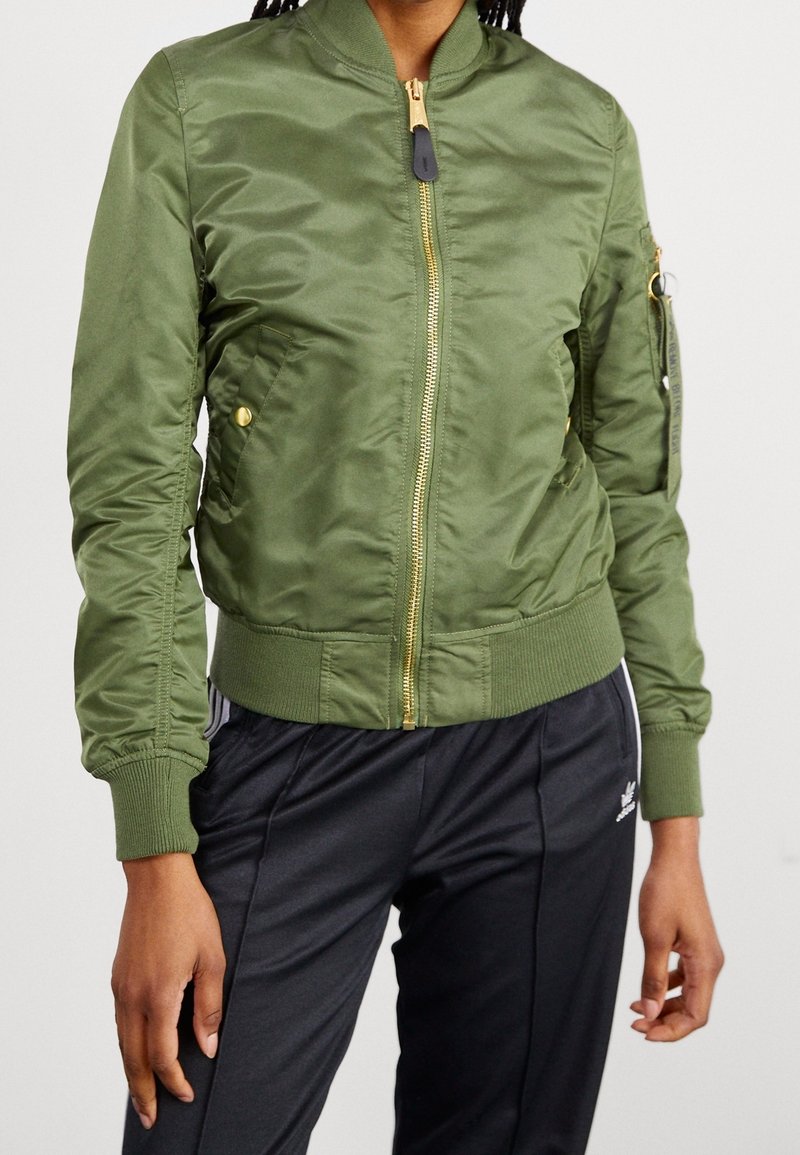 Personne portant une veste bomber verte ajustée avec poignets et taille côtelés, fermeture éclair dorée, et pantalon de survêtement noir avec des bandes blanches.
