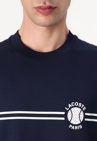 Námořnická modrá bavlněná tričko s kulatým výstřihem, se světlými vodorovnými pruhy a kruhovým logem Lacoste s textem "PARIS".
