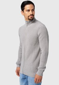 Suéter gris de canalé con cremallera y cuello alto, mangas ajustadas y tejido texturizado. Destacado con un sutil logotipo cerca del dobladillo.