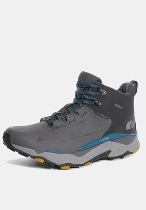 Bota de senderismo alta en gris y azul con suela resistente, acentos amarillos en la banda de rodadura, cuello acolchado y logo en el lateral que muestra "The North Face".