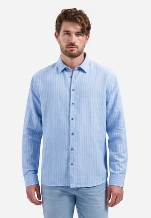 Mann mit lockigem Haar, der ein hellblaues, vertikal gestreiftes Hemd und hellblaue Jeans trägt, und vor einem weißen Hintergrund steht.