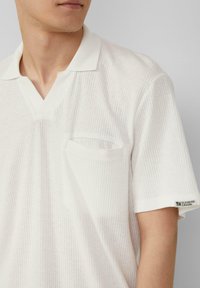 Witte, kortärmige polo met ribbelstructuur, V-hals en een enkele borstzak. Heeft een logo op de mouw.