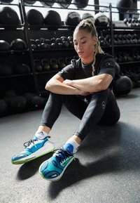 Modré a zelené sportovní boty s černými akcenty a strukturovaným svrškem, nošené s černými legínami a bílými ponožkami, sedící na podlaze v tělocvičně.