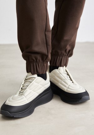 Person trägt weiße und schwarze Plateausneaker mit braunen Jogginghosen und steht auf einem hellgrauen Boden.