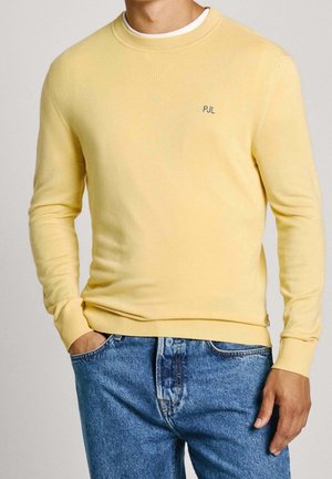Homme portant un pull jaune clair à col rond avec « PJL » sur la poitrine et un jean bleu, mains dans les poches.