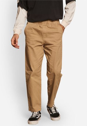 Homme portant un pantalon beige avec taille élastique et des baskets noires, main dans la poche, sur fond blanc uni.