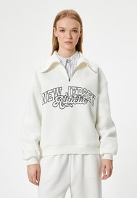 Biała bluza z wysokim kołnierzem, zapinana na zamek, z nadrukiem "NEW JERSEY Athletic" w kolorze czarnym. Miękka tkanina i luźny krój.
