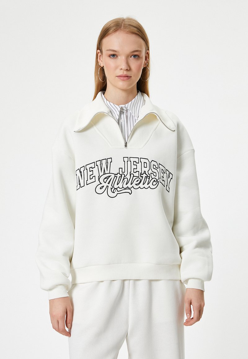 Hvid sweatshirt med høj krave, lynlås i halsen og "NEW JERSEY Athletic" trykt i sort. Blødt stof og afslappet pasform.