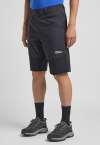 Pantaloni corti neri da esterno con multiple tasche con zip, realizzati in tessuto leggero. Indossati con calzini neri alla caviglia e scarpe da trekking grigie.