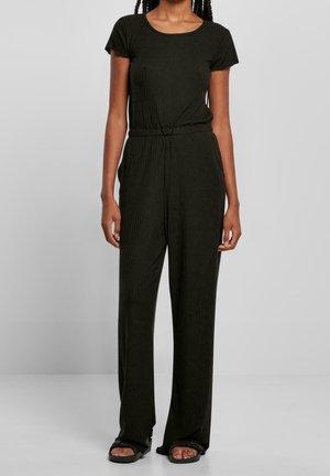 Kvinde iført en sort kortærmet ribbet jumpsuit med vidde bukser og sorte sandaler med åben tå, stående foran en ensfarvet baggrund.