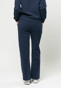 Navy krāsas augsto jostasvietu sporta bikses ar brīvu piegriezumu. Iespējas sānu kabatas un teksturēts audums, kombinētas ar baltām sporta kurpēm.