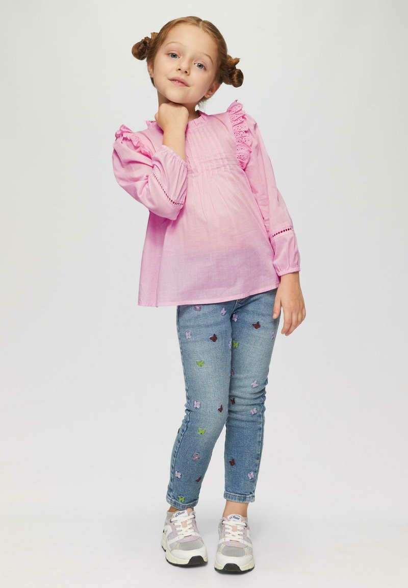 Lichtroze blouse met lange mouwen en ruffles, gecombineerd met blauwe jeans met multikleuren vlinder borduursels en witte sneakers.