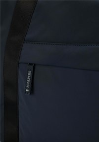 Bolsa de tela azul marino con textura suave, que cuenta con una correa negra y una etiqueta visible que dice "SCALPERS" en el bolsillo frontal.