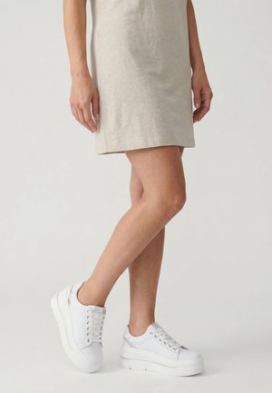 Vrouw die een beige jurk draagt en witte platformsneakers met zilveren accenten, staand tegen een effen lichtgrijze achtergrond.