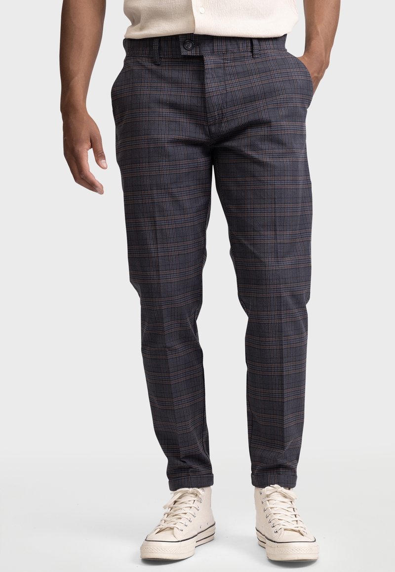 Homme portant un pantalon slim à carreaux foncé avec des poches avant, des baskets montantes blanches et une chemise beige rentrée.