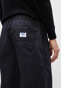 Calças cargo pretas com um bolso traseiro visível, apresentando um rótulo de marca azul e branco. Feitas de um material texturizado e durável.