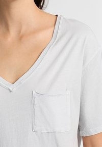 Svetle šedé tričko s krátkým rukávem a výstřihem do V, malou náprsní kapsou a viditelnými švy podél výstřihu.
