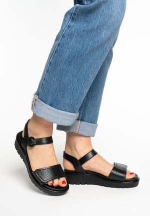 Feet wearing black textured leather sandals with straps and buckles, paired with cuffed blue denim jeans against a white background.