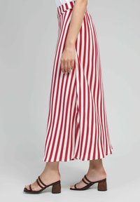 Jupe longue et fluide avec des rayures verticales rouges et blanches. Fabriquée en tissu léger, elle a une taille haute et un ourlet modeste. Portée avec des sandales à talons carrés marron.