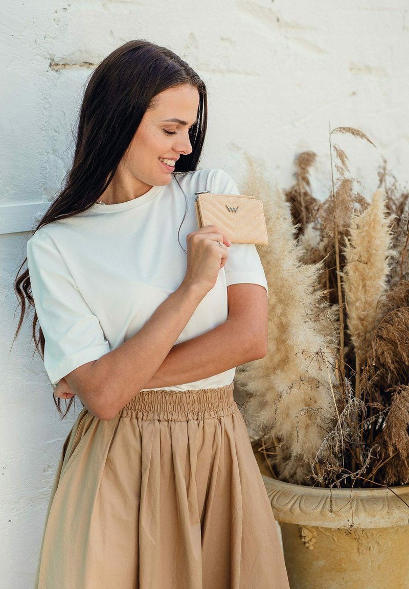 Beige peněženka na zip s chevron vzorem a logem, kterou drží žena v bílém topu a béžové sukni, poblíž dekorativních sušených rostlin.