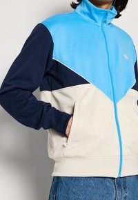 Träningsjacka i blått, ljusblått, marinblått och creme; har dragkedja fram, fickor och kontrasterande färgblock med logotyp.