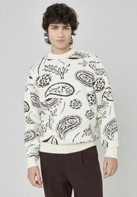 Pull blanc avec un motif paisley et floral brun, avec des poignets et un ourlet côtelés. Modèle associé à un pantalon brun foncé, se tenant devant un fond clair.
