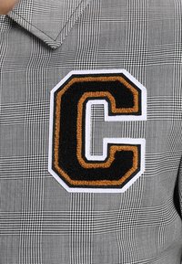 Chemise en tissu à carreaux noirs et blancs, avec un patch "C" texturé en noir et or, brodé en relief, sur le côté gauche.