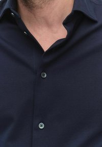 Marineblauwe button-up overhemd met een textuurstof, twee zichtbare knopen en een gespreide kraag. Close-up opname van het nekgebied.