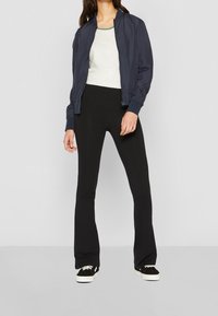 Merinotakk med marineblått fôr over en hvit T-skjorte, kombinert med svarte vide bukser og svarte sneakers med hvite såler. Stoffet ser glatt og elastisk ut.